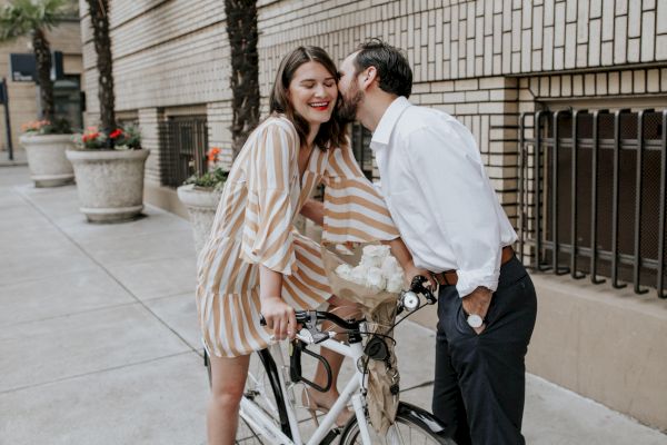 A couple enjoys a moment by a bike on a sidewalk, with a building and palm trees in the background, as one leans to kiss the other's cheek.