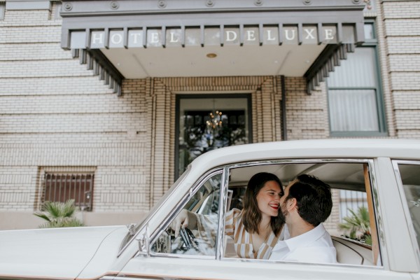 A couple shares a kiss inside a classic car parked in front of a building with a sign reading 