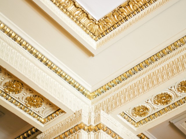 The image shows an ornate interior ceiling corner with detailed gold and white molding and decorative elements.