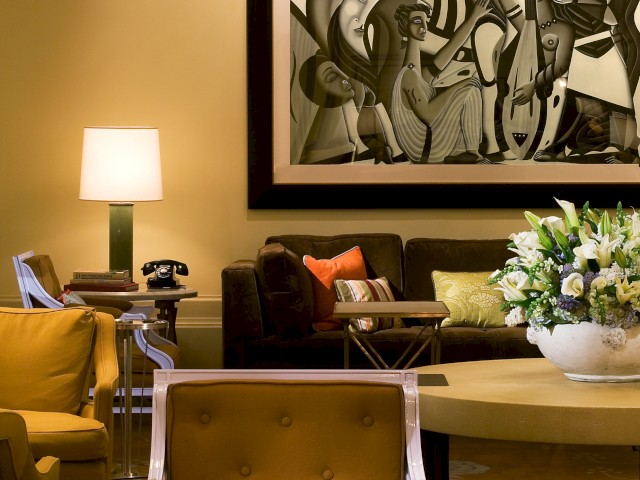 A modern living room with yellow chairs, a lamp, a large abstract painting, a chandelier, and a round table with a flower arrangement.