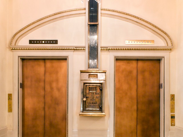 The image shows two brass elevator doors inside a luxurious lobby with a chandelier above and an old-fashioned mail chute in the center of the wall.