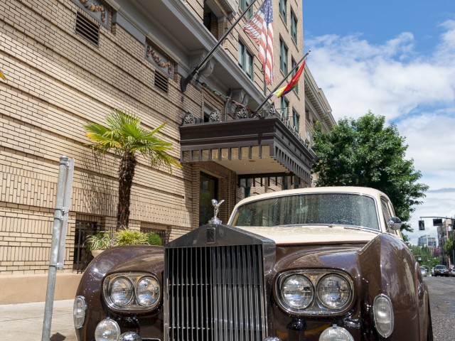 A classic Rolls-Royce is parked in front of a building with American and California flags. License plate reads 