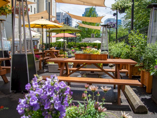 An outdoor patio with wooden picnic tables, colorful flowers, umbrellas providing shade, and greenery in the background.