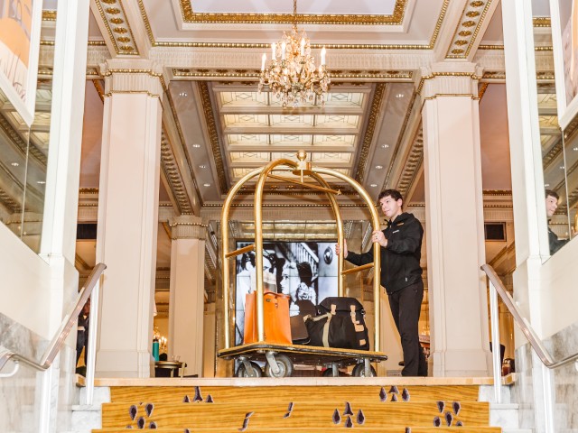 A hotel bellhop stands on a staircase next to a loaded luggage cart in a luxurious lobby with chandeliers and ornate decorations.