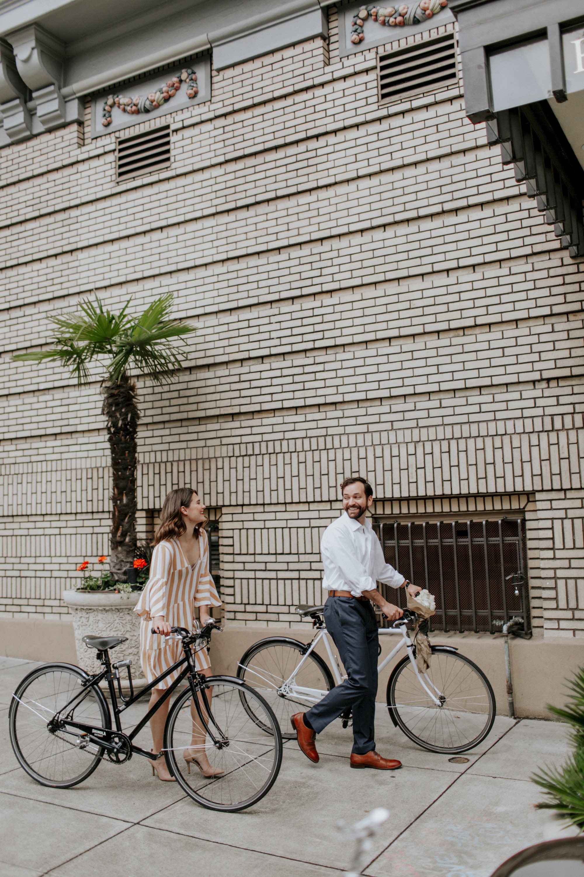 A man and a woman are walking with their bicycles along a street with a brick wall and a small palm tree in the background.