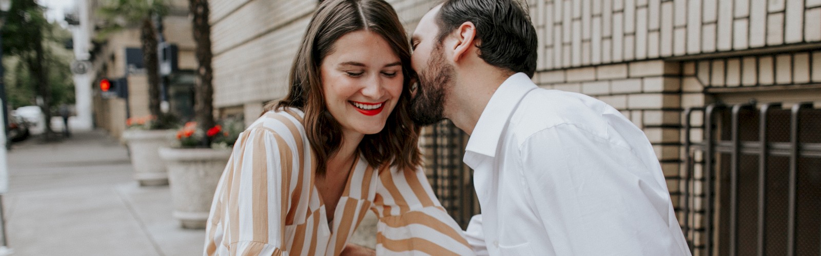 A couple is on a street; the woman is on a bicycle with flowers, and the man leans in to whisper or kiss her cheek, both smiling.