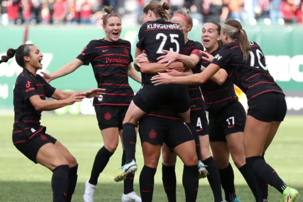 A group of female soccer players wearing black uniforms are celebrating on the field, with one player being lifted by her teammates.