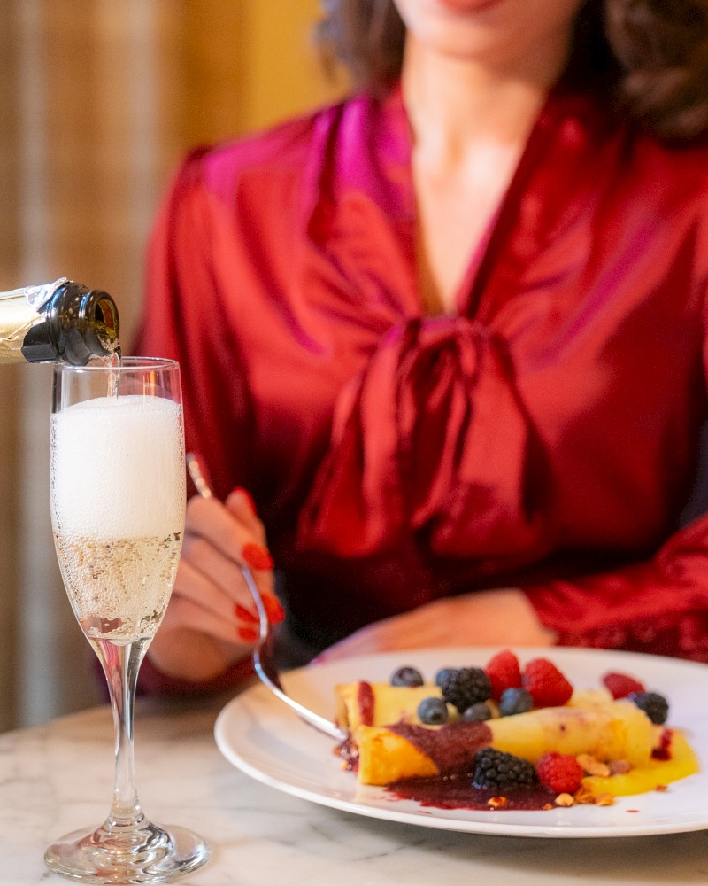 A person in a red blouse is seated, holding a fork with a dessert plate and berries. Another person is pouring champagne into a glass.