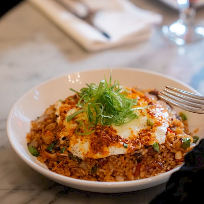 The image shows a bowl of fried rice topped with a fried egg and garnished with green onions, placed on a marble table next to cutlery and a napkin.