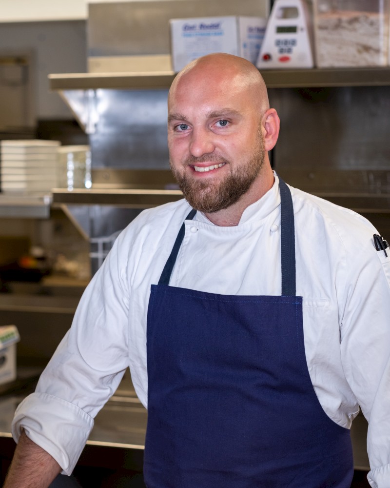 A man wearing a white chef's coat and a blue apron is smiling while standing in a kitchen with stainless steel appliances.