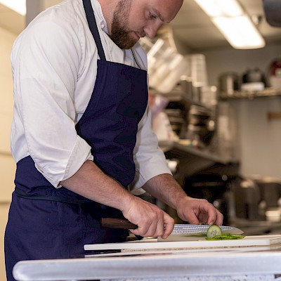 A person in a kitchen is cutting a vegetable on a white cutting board while wearing a white shirt and dark blue apron.