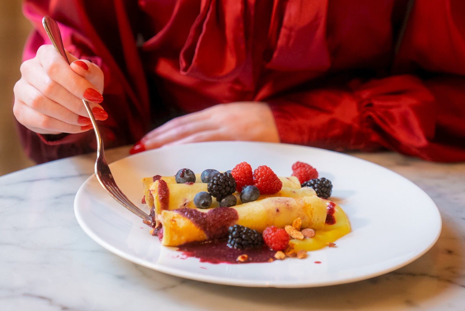 A person in a red blouse is about to eat a dessert crepe topped with various berries on a white plate at a marble table.