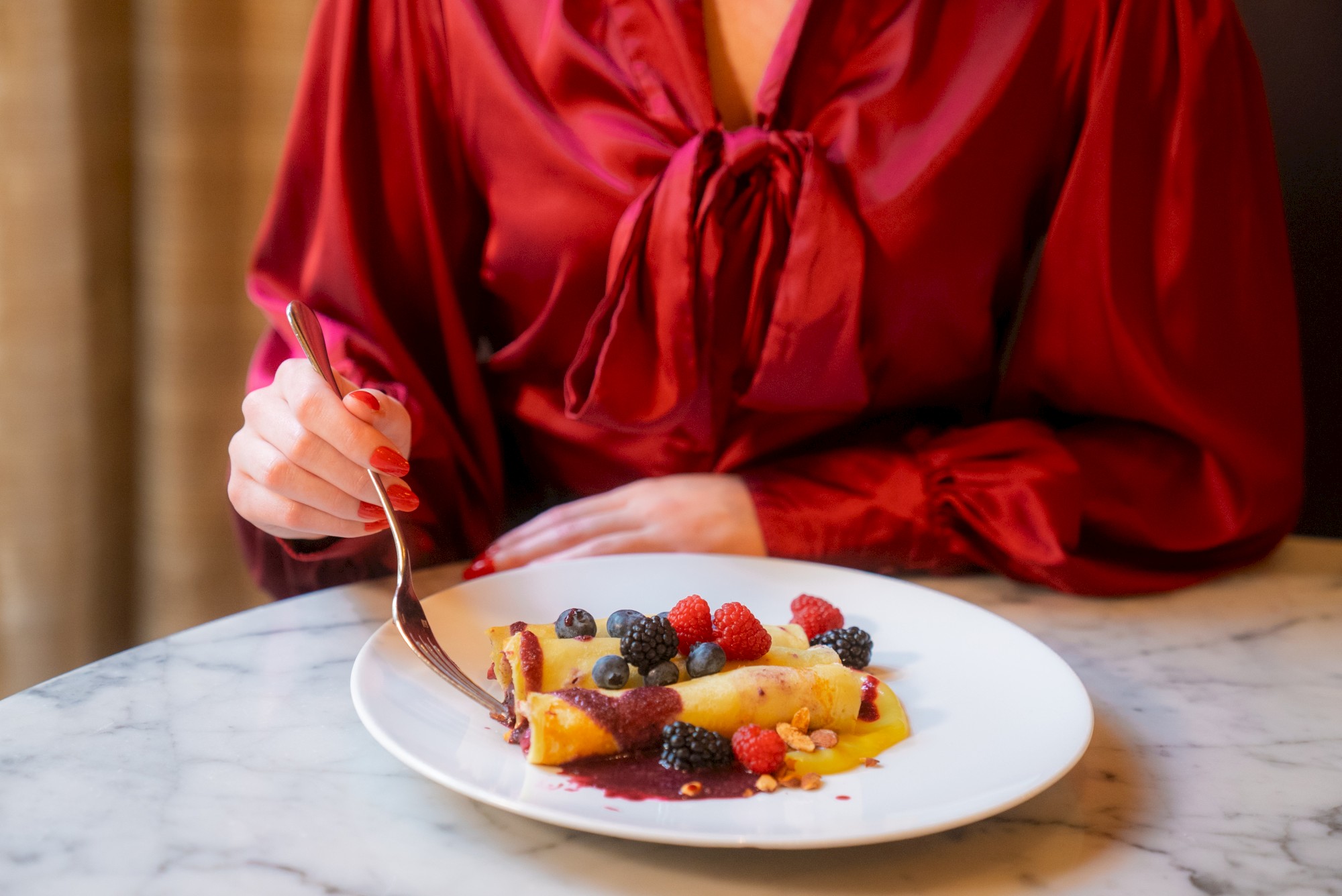 A person in a red blouse is about to eat a dessert crepe topped with various berries on a white plate at a marble table.