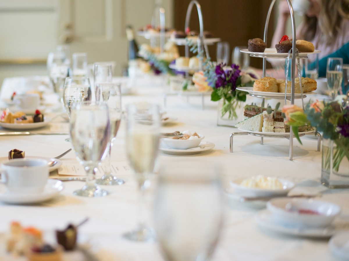 A well-set table with tiered trays of pastries and desserts, glasses, teacups, and floral centerpieces in a banquet or high tea setting.