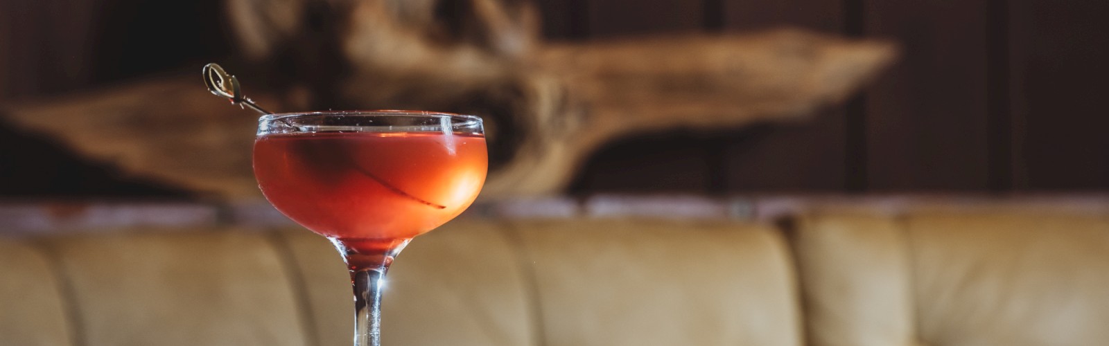 A cocktail in a stemmed glass sits on a dark table with a wooden background and a cushioned bench.