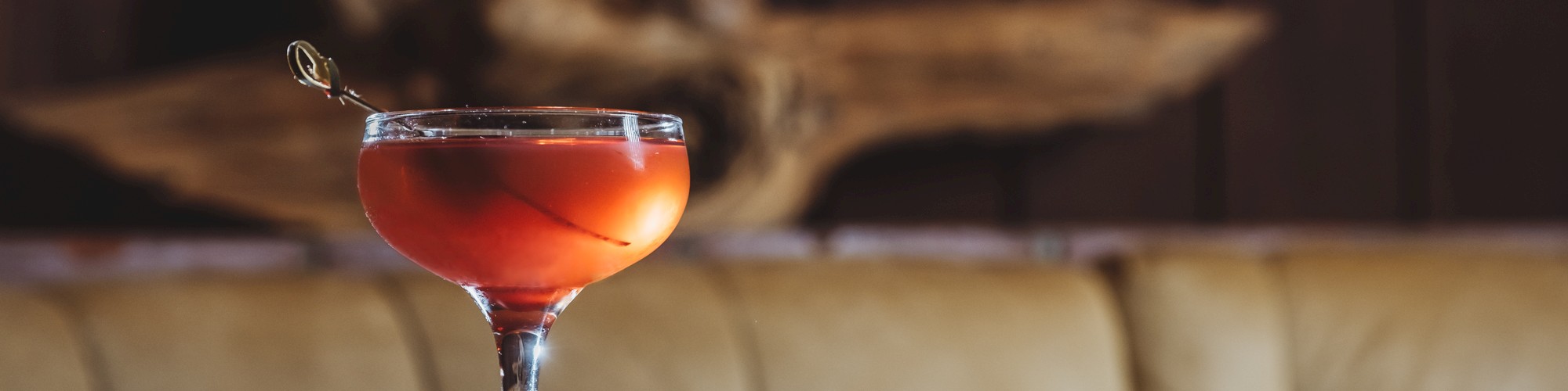 A cocktail in a stemmed glass sits on a dark table with a wooden background and a cushioned bench.