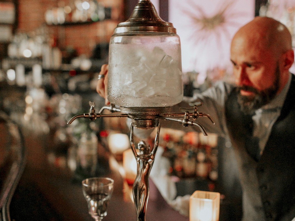 A bartender behind a counter in a dimly lit bar, with an ornate absinthe fountain and glasses on the counter, along with a few candles.