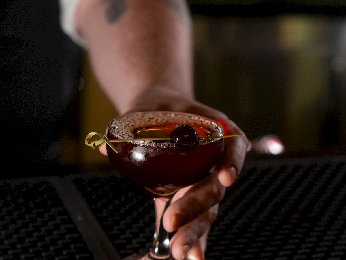 A person holding a cocktail glass filled with a dark-colored drink, with a cherry garnish on top, in a dimly lit bar setting.