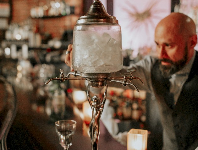 A bartender is preparing a drink using an ornate absinthe fountain on a bar counter, with candles and shelves of bottles visible in the background.