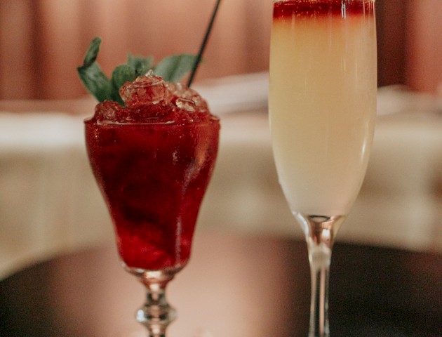 Two cocktails on a table; one is red with mint leaves and ice, while the other is a yellow drink topped with a red layer.