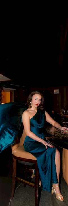 Woman in a blue dress sitting at a bar, with flowing fabric behind her, in a dimly lit lounge with bar stools and drinks visible.