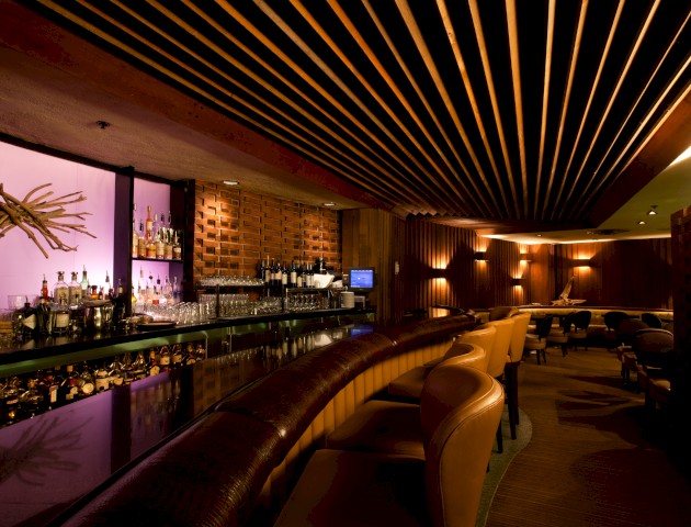 This image shows a modern bar with a curved counter, leather stools, an extensive selection of bottles, dim lighting, and cozy seating areas.