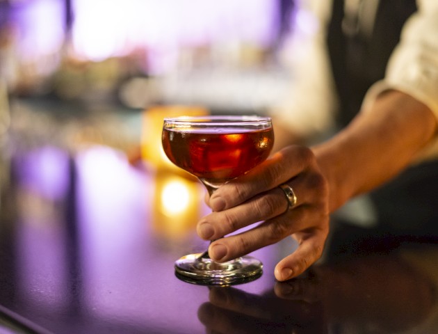 A person’s hand holding a cocktail glass filled with a dark-colored drink, set against a blurred background of a bar.