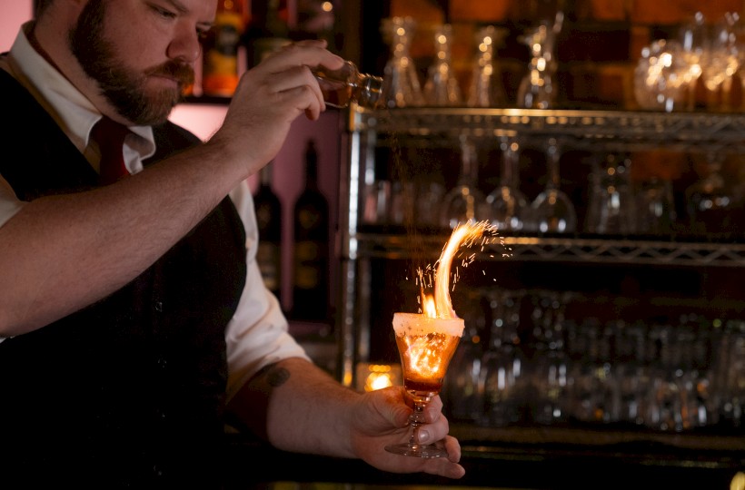 A man is igniting a drink in a dimly lit bar, creating a burst of flame from the cocktail he is holding.