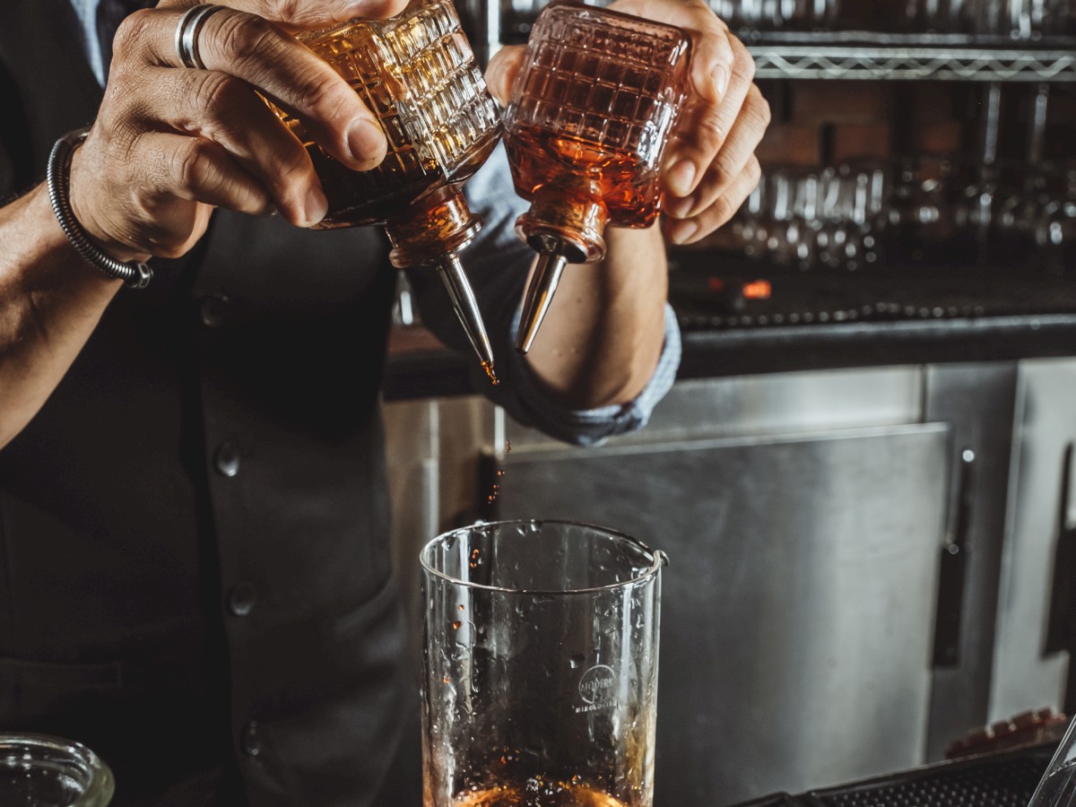 A bartender pouring drinks into a mixing glass with both hands in a bar setting, with shelves of glasses and bar equipment in the background.