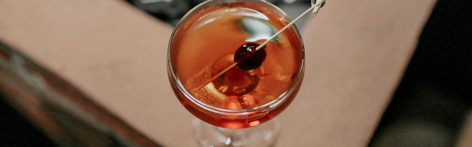 A top view of a cocktail in a glass, garnished with a cherry on a cocktail pick, positioned on a stone and wood table.