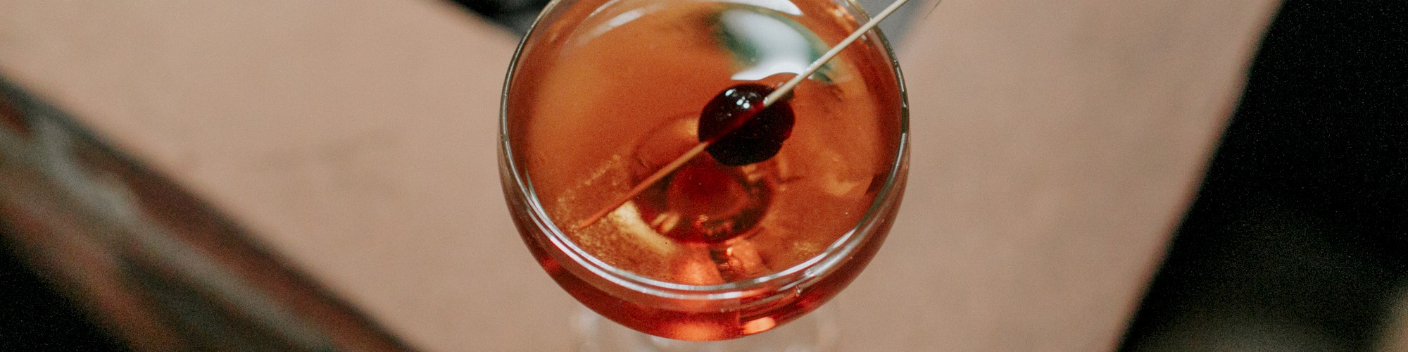 A top view of a cocktail in a glass, garnished with a cherry on a cocktail pick, positioned on a stone and wood table.