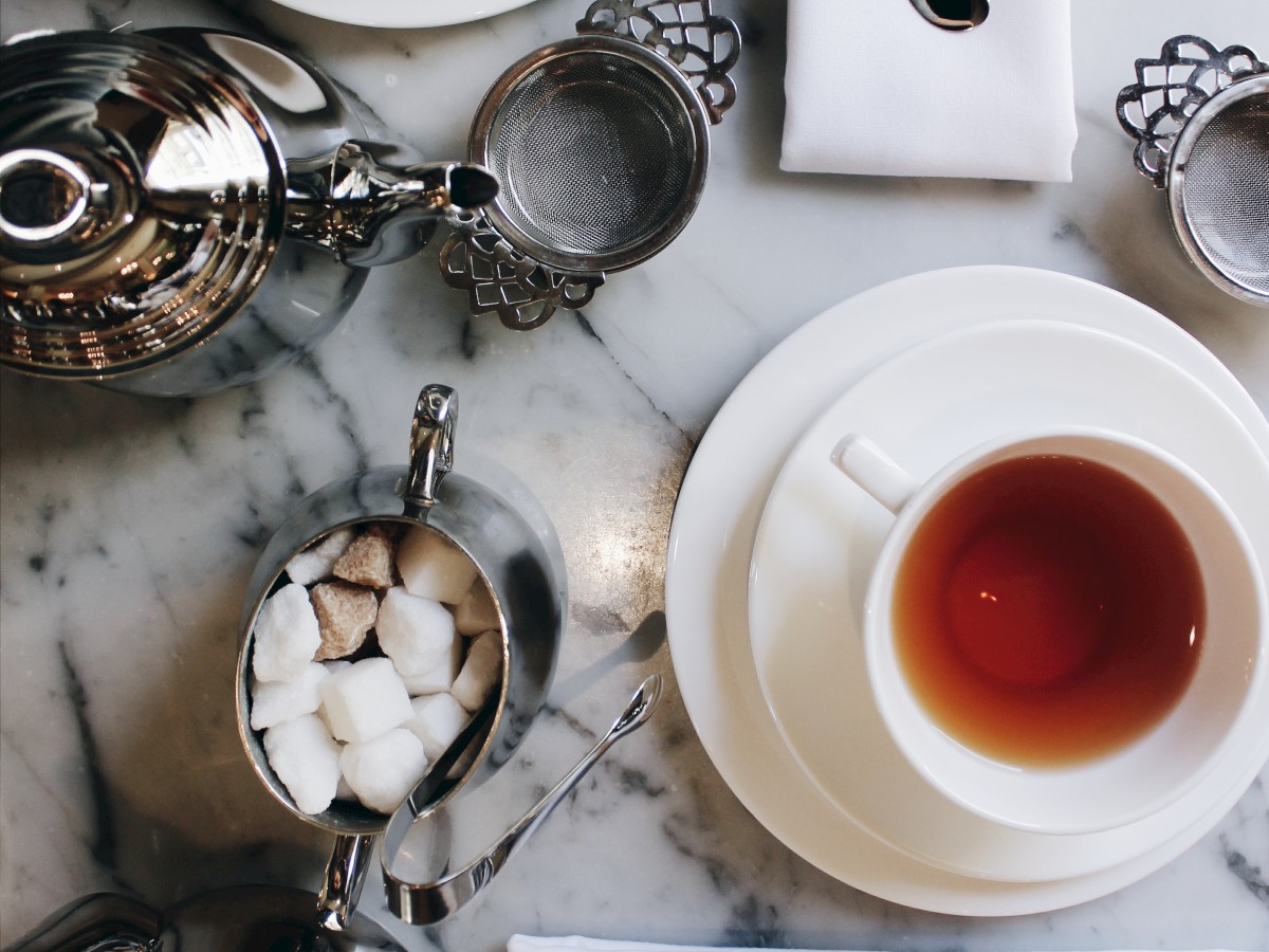 The image shows a tea setup with two cups of tea, teapots, sugar cubes, and neatly arranged cutlery on a marble table.