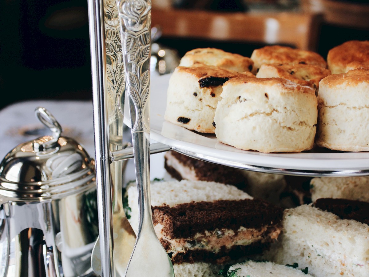 A tiered tray with assorted pastries, scones, and sandwiches on a table, alongside a silver teapot, ready for afternoon tea.