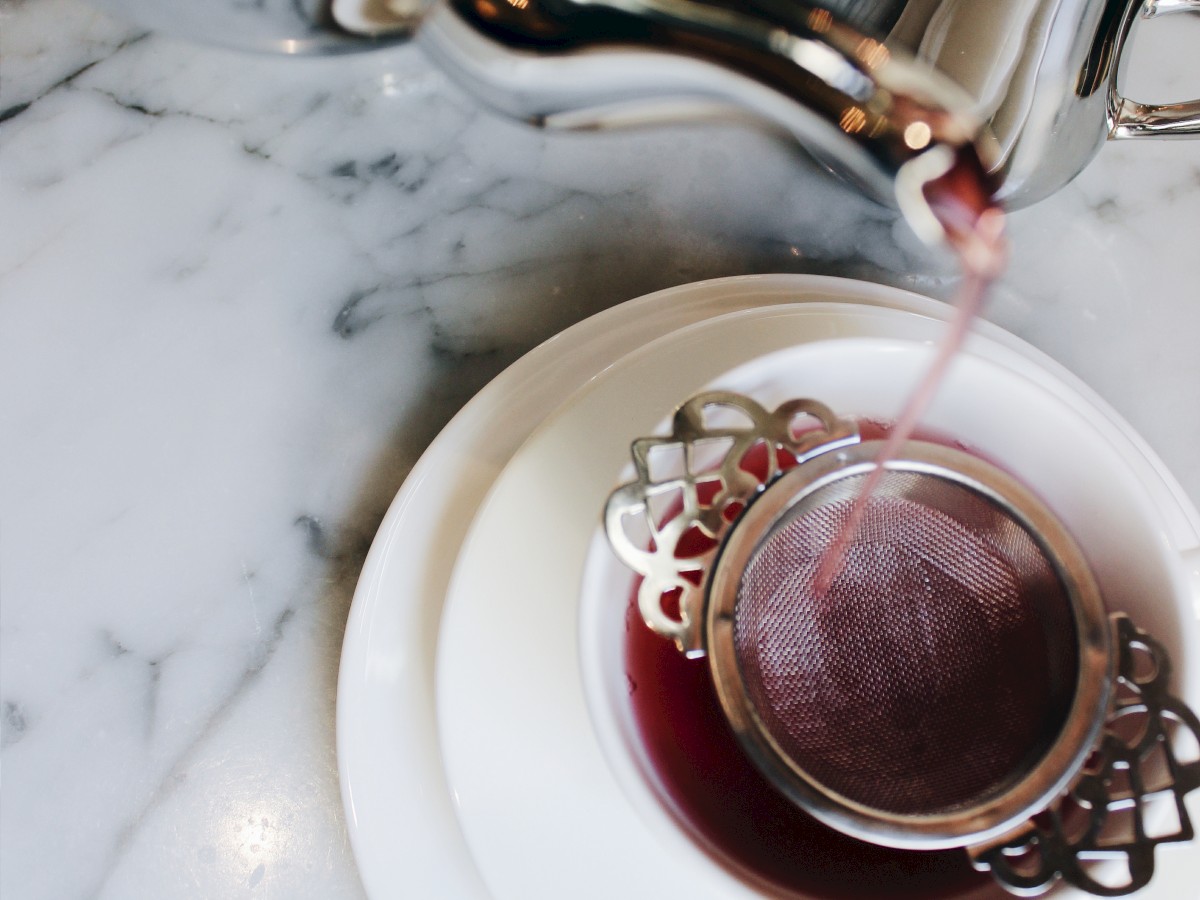 Tea is being poured from a teapot into a cup with a metal strainer, on a marble surface.