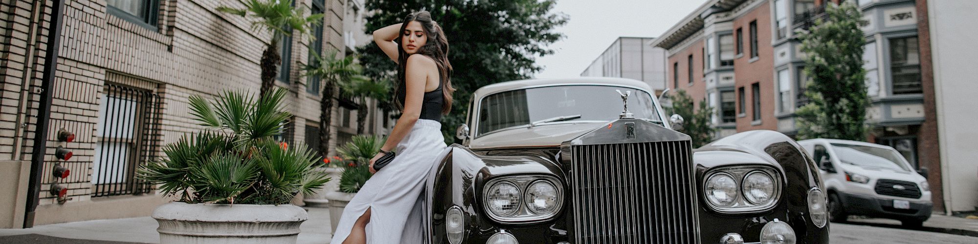 A woman in a white dress poses next to a classic car on a city street, with various buildings and a potted plant visible in the background.