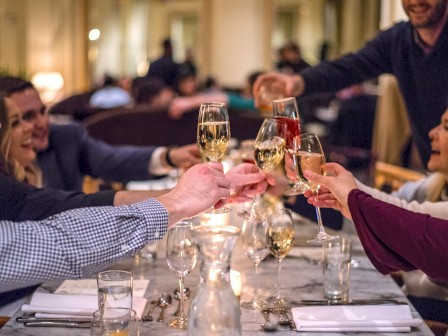 A group of people sitting around a table in a restaurant are clinking glasses in a celebratory toast, creating a lively and cheerful atmosphere.