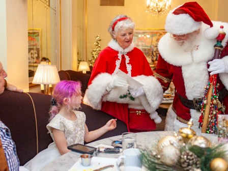 Santa and Mrs. Claus visit a family in a festive setting, with a girl wearing pink highlighting the joyful scene.