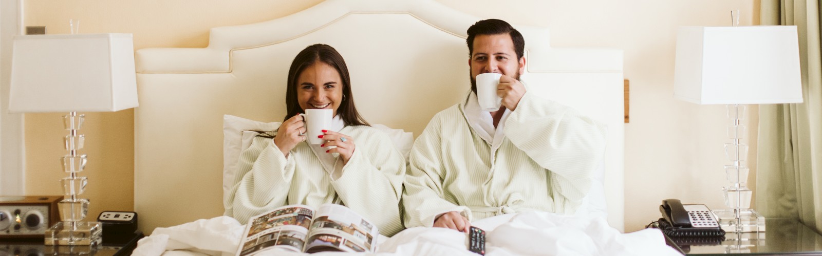 A couple in robes sits in bed, sipping from mugs with a magazine open. Two lamps and a framed picture adorn the room, creating a cozy ambiance.