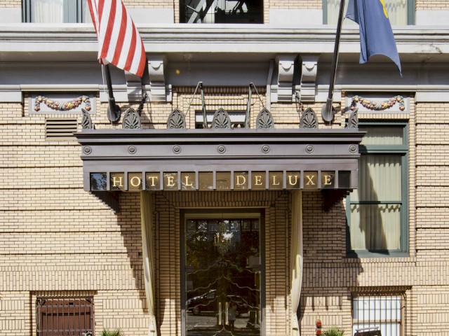 The image shows the entrance of Hotel DeLuxe with American and state flags, set against a brick facade adorned with plants.
