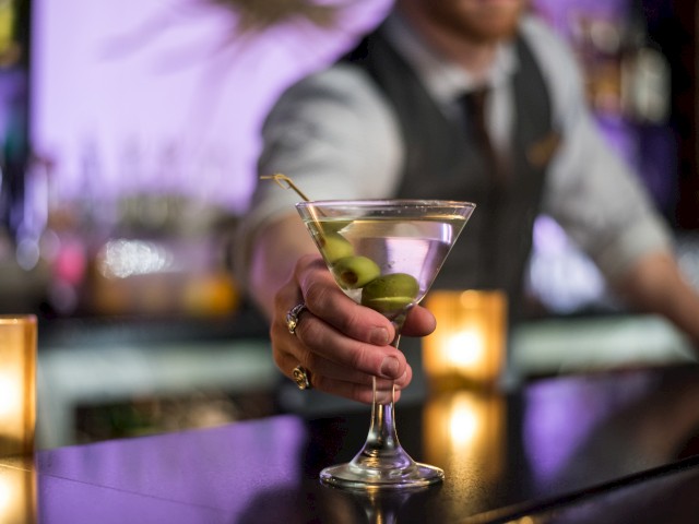 A bartender is serving a martini with olives at a bar, with a blurred background and warm lighting in the scene.