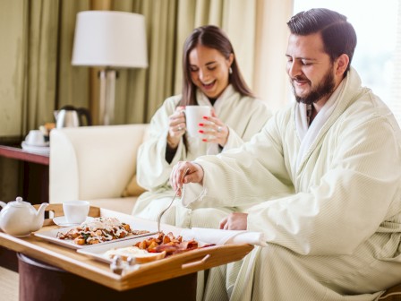 A man and woman in robes enjoy breakfast in a cozy living room. They have a tray with food and drinks on a table, smiling and relaxed.