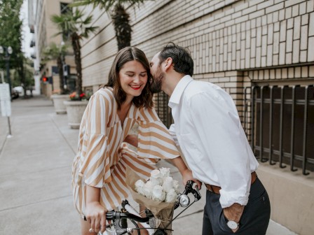 A woman on a bike with flowers smiles as a man leans in to speak to her on a city sidewalk.