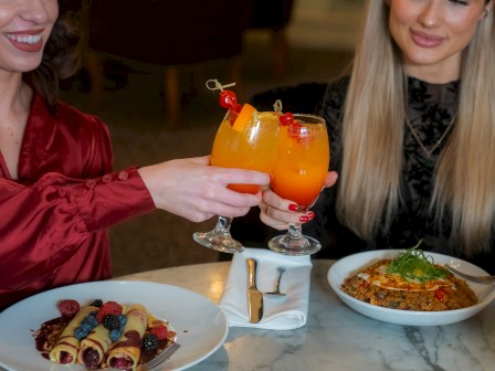 Two people at a table with colorful drinks, berry-filled crepes, and a quinoa dish, ready to enjoy a meal together.