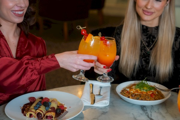 Two people at a table with colorful drinks, berry-filled crepes, and a quinoa dish, ready to enjoy a meal together.