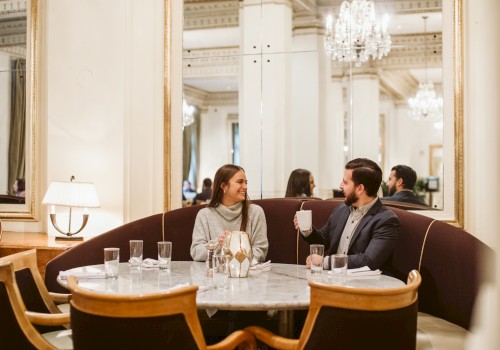 A man and woman are sitting at a round table in a stylish restaurant with elegant decor and chandeliers, engaging in conversation.