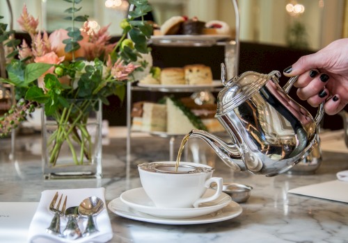 A hand pours tea from a silver teapot into a cup on a marble table with flowers and a tiered tray of pastries in the background.