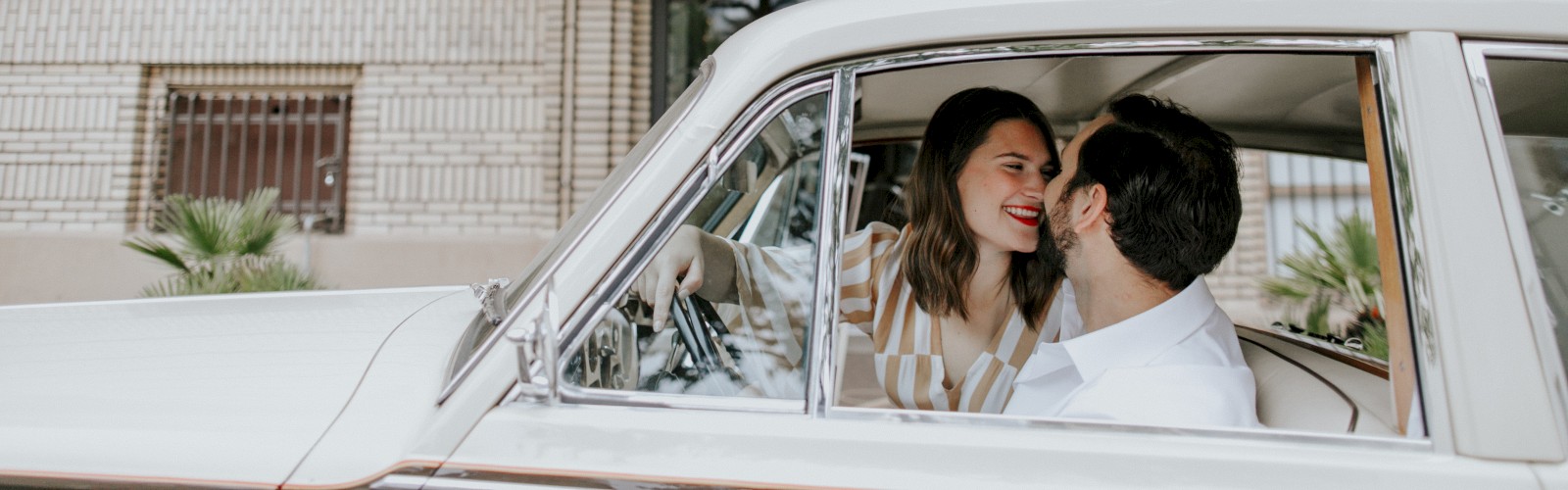 A couple is sitting in a vintage car parked outside a building with a 