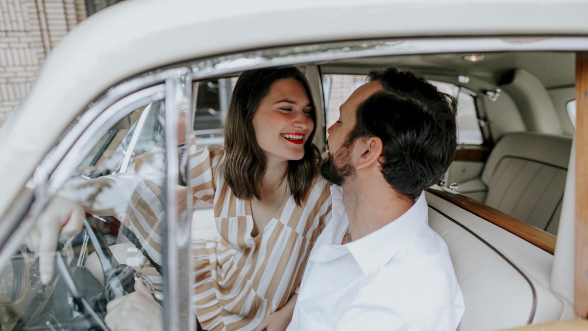 A couple is sitting inside a classic car, smiling at each other. The woman leans in toward the man, creating a happy and intimate moment.