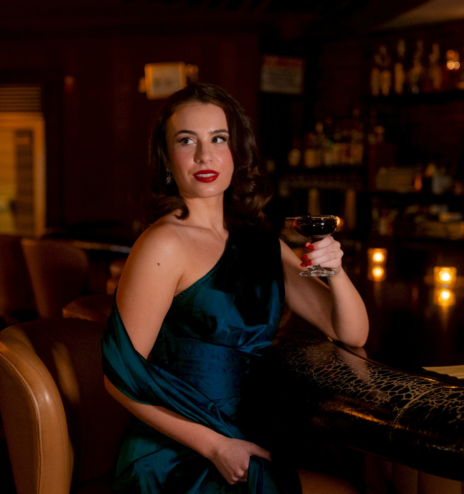 A woman in a blue dress at a bar, holding a glass of wine, in a dimly lit setting.