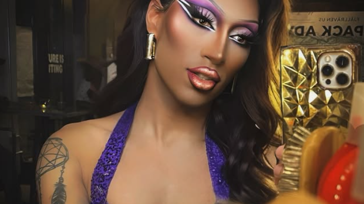 A person in glamorous makeup wearing a purple outfit takes a selfie in a mirror, surrounded by beauty and decor items.